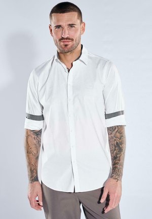 Mann mit kurzen Haaren und Tätowierungen steht vor schlichtem Hintergrund, trägt ein weißes Hemd mit aufgerollten Ärmeln und graue Hose.