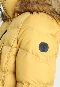 Gelbe Pufferjacke mit gefütterter Kapuze aus Kunstfell, genähtem Muster, seitlichen Taschen und einem schwarzen Logo-Patch auf dem oberen Ärmel.