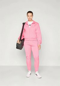 Ροζ αθλητική φόρμα με ζακέτα που κλείνει με φερμουάρ και εφαρμοστά παντελόνια, συμπληρωμένη από μια μαύρη τσάντα ώμου και λευκά αθλητικά παπούτσια με μαύρες λεπτομέρειες.