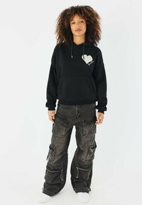 Felpa nera con cappuccio decorata con una grafica a forma di cuore, abbinata a pantaloni cargo neri larghi e texturizzati con molteplici tasche e cerniere. Sneakers bianche.
