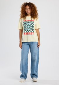 Yellow graphic t-shirt featuring a fish pattern and "SEAFOOD MARKET" text in red, paired with light-blue wide-leg jeans and white sneakers.