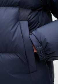 Ναυτικό μπλε φουσκωτό μπουφάν με γυαλιστερή, λεία υφή. Διαθέτει πλαϊνή τσέπη και ελαστικούς μανικιών για άνετη εφαρμογή.