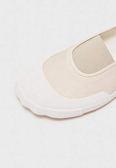 Beige Slipper mit einer strukturierten weißen Gummisohle, ausgestattet mit einem glatten Obermaterial und einem elastischen Band für einfaches Anziehen. Mit gewelltem Zehendesign.
