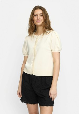 Cremefarbener kurzärmeliger Cardigan mit Rundhalsausschnitt und fünf Knöpfen, kombiniert mit schwarzen bestickten Shorts mit gewelltem Saum.