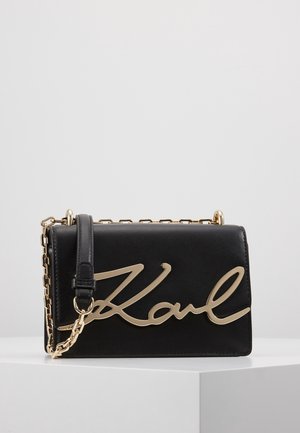 Sac à main en cuir noir avec une bandoulière chaîne dorée et une grande signature "Karl" dorée sur le devant, posé sur une surface blanche.