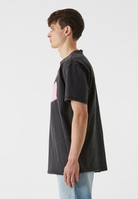 Melna kokvilnas pārmērīga t-kreklu ar īsām piedurknēm, ar lielu rozā grafiku priekšpusē. Sānu skats parāda brīvu piegriezumu un apaļu kakla izgriezumu.