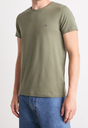 Homme portant un t-shirt manches courtes vert olive avec un petit logo rectangulaire sur la poitrine et un jean denim bleu sur un fond uni.