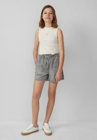 Lichtbeige mouwloze top met een klein oranje graphic, gecombineerd met grijze shorts met een elastische tailleband en zijzakken. Witte sneakers.