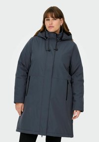 Sieviete, kurai mugurā tumši pelēka ziemas jaka ar kapuci un rāvējslēdzēja kabatām, stāv vienkārša gaiša fona priekšā.