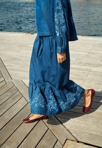 Denim obleka z dolgimi rokavi, okrašena s svetlo modrim cvetličnim vezenjem. Rob ima lataste detajle. Oblečena z burgundskimi balerinkami na lesenih desnah.