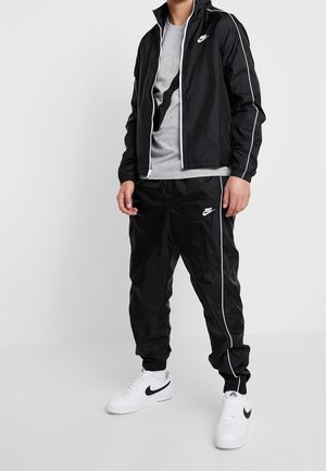 Jong persoon draagt een zwart Nike trainingspak met witte biezen, een grijs T-shirt met zwart logo en witte Nike sneakers, staand tegen een witte achtergrond.