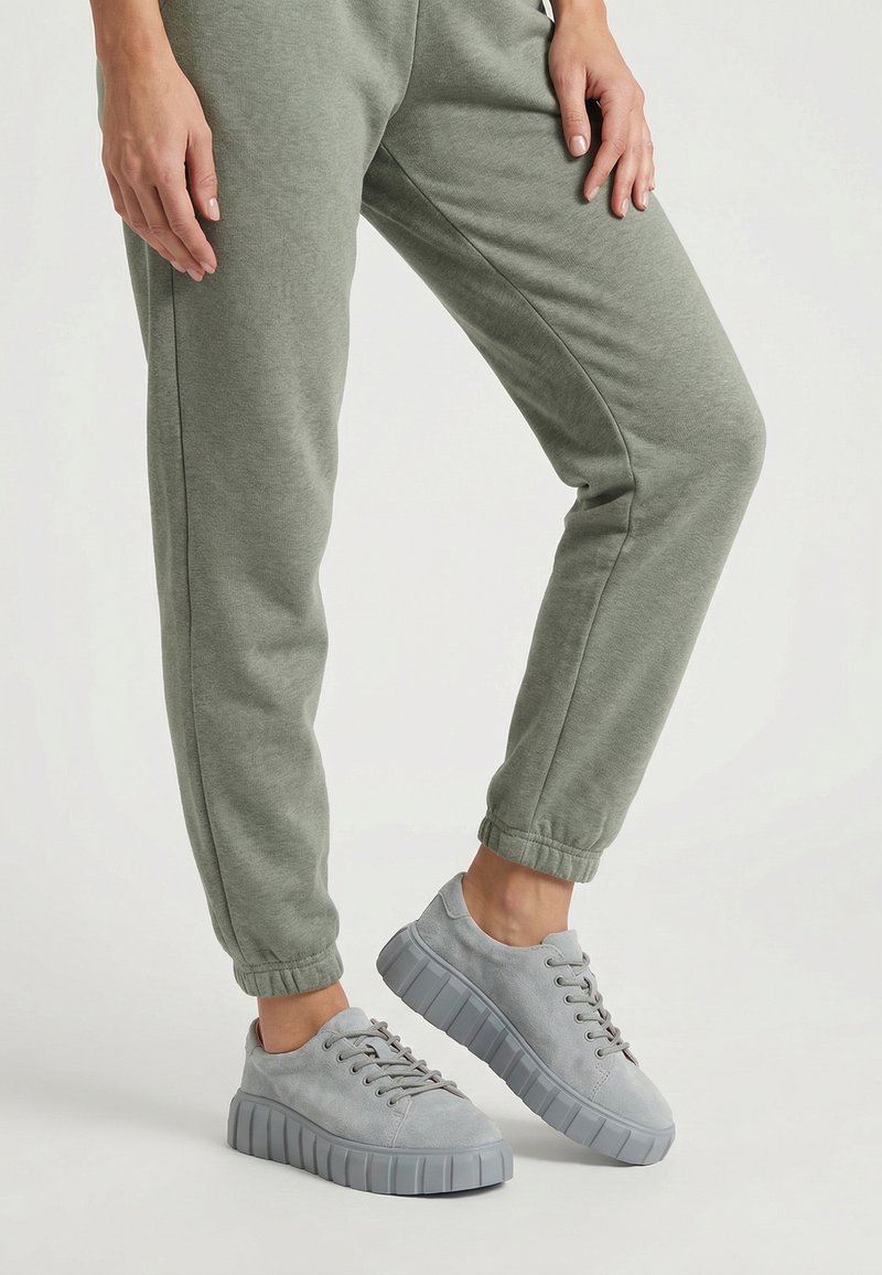 Osoba nosząca jasnoszare tenisówki z grubymi podeszwami oraz zielone spodnie dresowe ze ściągaczami, stojąca z jedną lekko uniesioną nogą.