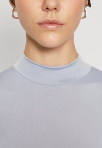 Lyseblå, høyhalset topp laget av et mykt og glatt stoff. Har en tettsittende design og minimale sømdetaljer. Metallring øredobb synlig.