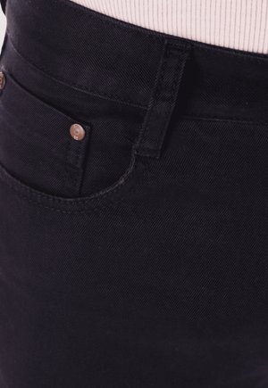 Los pantalones vaqueros de mezclilla negra presentan un diseño clásico de cinco bolsillos con cierre de cremallera y botón. Se pueden apreciar costuras sutiles y detalles metálicos.