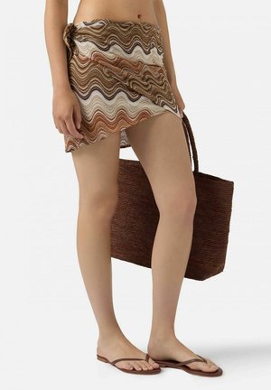 Donna che indossa una gonna sarong marrone con motivo ondulato, infradito marroni, che tiene in mano una grande borsa tote intrecciata marrone.