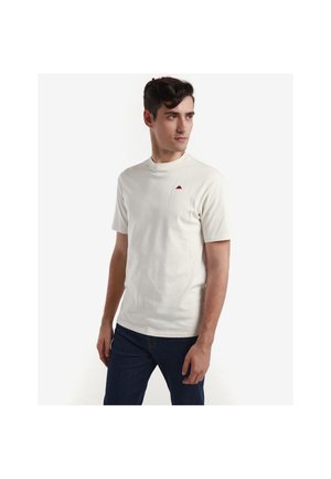 Hombre con una camiseta de manga corta lisa color blanco roto con un pequeño logo rojo en el pecho izquierdo y vaqueros azul oscuro, mirando hacia la derecha.