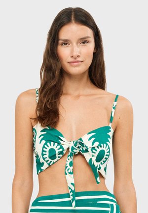 Joven con cabello largo y castaño, vistiendo un top corto anudado con estampado verde y blanco y parte inferior a rayas verdes, sobre un fondo liso.