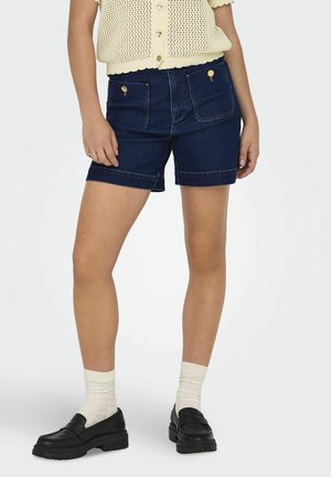 Donkere blauwe denimshorts met een hoge taille, voorzien van gouden hardware en achterzakken. Gepaard met zwarte instappers en crèmekleurige sokken.