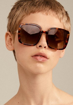 Occhiali da sole tartaruga con montatura quadrata oversize, lenti scure sfumate e dettagli dorati sulle tempie. Materiale traslucido e finitura lucida.