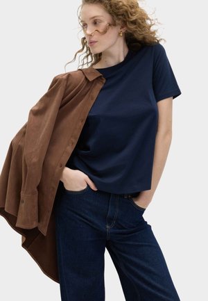 Vrouw met krullend haar, gekleed in een marineblauw T-shirt en een donkere spijkerbroek, met een bruin jasje over één schouder, kijkt opzij.