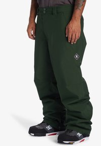 Green snow pants with a smooth texture, featuring two front buttons, side pockets, and a diamond logo on the right thigh.