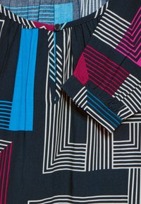 Långärmad topp i marinblått med flerfärgade geometriska mönster. Har en knappstängning vid halsen och randiga detaljer på ärmsluten.