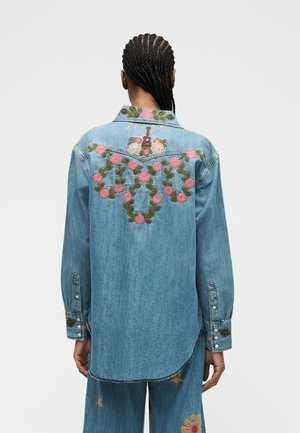 Person mit geflochtenem Haar, die ein hellblaues Jeanshemd trägt, das auf der Rückseite mit bestickten pinken Blumen, grünen Blättern und einer kleinen braunen Gitarre verziert ist.