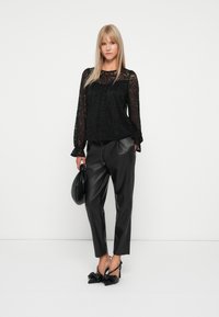 Zwarte kanten blouse met lange mouwen en gerimpelde manchetten, gecombineerd met hoge zwarte leren broek van kunstleder. Accessoires zijn een zwarte tas en hakken.