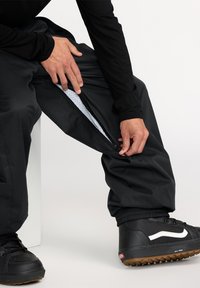 Svarta vattentäta byxor med zippered sidöppning, med nätfoder och elastiska ärmar, i kombination med svarta sneakers.