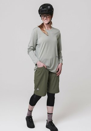 Frau mit schwarzem Helm, Brille, hellgrauem Langarmshirt, olivgrünen Shorts, schwarzen Knieschonern und schwarzen Schuhen, die mit den Händen in den Taschen steht.