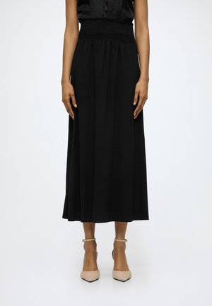 Femme portant une jupe midi noire avec taille haute smockée et talons beiges à bride à la cheville et bout pointu, debout sur fond blanc.