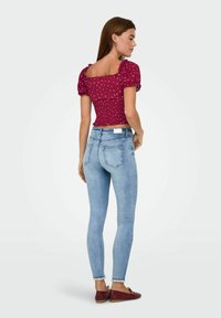 Eine Frau trägt ein burgunderfarbenes gepunktetes Oberteil mit Puffärmeln, helle blaue Skinny Jeans und burgunderfarbene Ballerinas. Sie steht mit dem Rücken zur Kamera und hat den Kopf nach rechts gedreht.