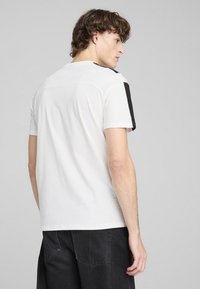Camiseta de algodón blanca con mangas cortas, cuello redondo y detalles en negro en los hombros. Textura suave, ajuste casual.