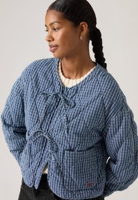 Γυναίκα με πλεξούδες φοράει ένα μπλε και λευκό καρό μπουφάν με επένδυση, δεμένο με φιόγκους, στέκεται με τα χέρια στις τσέπες, κοιτώντας προς τα πλάγια.