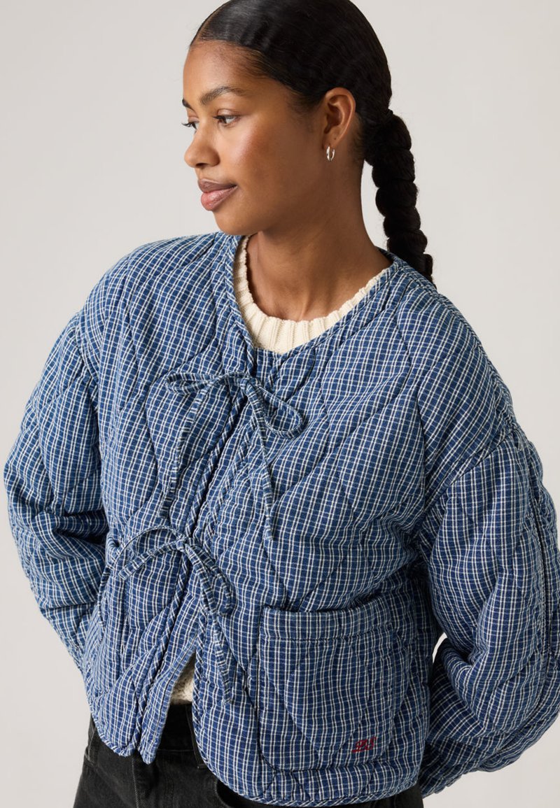Femme aux cheveux tressés portant une veste matelassée à carreaux bleue et blanche nouée avec des nœuds, debout les mains dans les poches, regardant sur le côté.