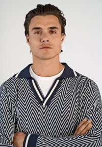 Pullover lavorato a maglia in un motivo a spina di pesce blu navy e bianco, con scollo a V profondo, ampio colletto e polsini a strisce contrastanti.