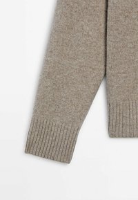 Extrémités des manches d'un pull beige en tricot avec des poignets côtelés, présenté sur un fond blanc uni.