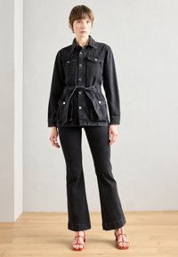 Black denim jacket with front pockets and waist tie, paired with black flared jeans. Red strappy sandals add color contrast.