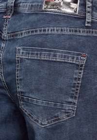 Jeans de mezclilla azul oscuro con un bolsillo trasero. Costuras visibles en blanco, una etiqueta en la cintura y una textura ligeramente desgastada.