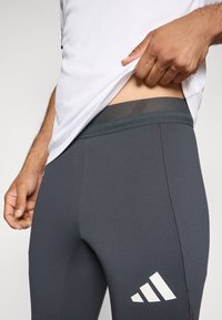 Leggings de sport en tissu gris foncé, avec une taille en mesh et un logo blanc bien visible. Texture lisse et design ajusté.