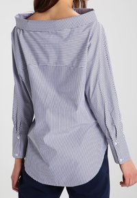 Camicia a righe blu navy e bianca, con un ampio colletto, maniche lunghe e orlo curvo. Il tessuto ha una texture liscia con sottili righe.