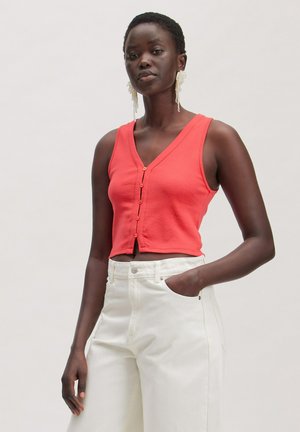 Vrouw draagt een mouwloos rood cropped topje met knopen en witte hoge taille broek, staat met één hand in de broekzak tegen een effen achtergrond.
