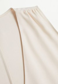 Zachte, rekbare stof in lichtbeige met een diepe V-halsontwerp; bevat geplooide details op de schouders voor extra textuur.