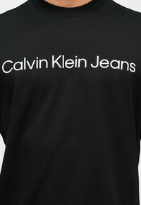 Μαύρο μπλουζάκι με στρογγυλή λαιμόκοψη και την επιγραφή «Calvin Klein Jeans» σε λευκό, τοποθετημένη στο κέντρο του στήθους.