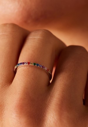 Regenboog edelstenen ring met een dunne metalen band, voorzien van meerkleurige stenen die langs de bovenrand zijn uitgelijnd tegen een hand.