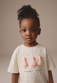 Lyse beige t-shirt med korte ærmer og ternet mønster, dekoreret med tre pink broderede ænder og teksten "ALWAYS CURIOUS" nedenunder.