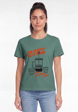 Femme aux cheveux attachés portant un t-shirt vert avec une illustration de cafetière à piston et des tasses, ainsi que le texte "Coffee makes everything better."