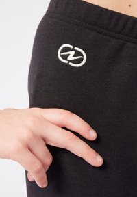 Pantaloni felpati neri in cotone con logo bianco ricamato. Il tessuto è liscio e morbido con una vita elastica. Una mano tocca delicatamente la gamba.