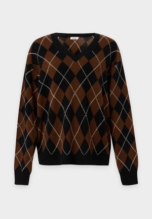 Femme portant un pull à losanges marron et noir, un jean bleu clair, des mocassins marron et tenant un sac à main marron devant un fond uni.