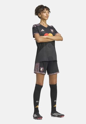 Vrouw staat zelfverzekerd met armen over elkaar, gekleed in een zwart voetbaluniform, kniekousen en voetbalschoenen tegen een effen lichte achtergrond.